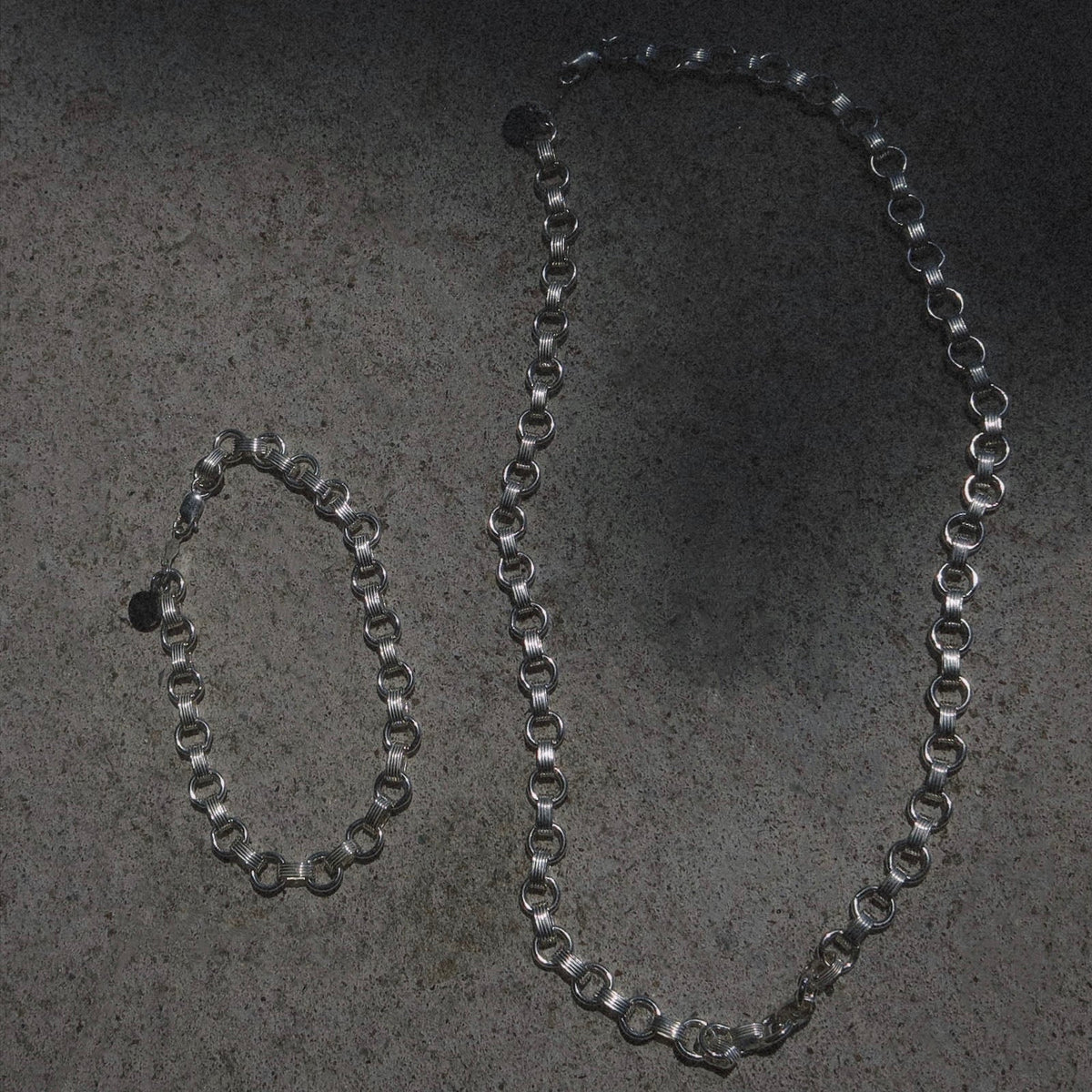 A silver chain necklace and matching bracelet are laid out on a textured gray surface, casting faint shadows.