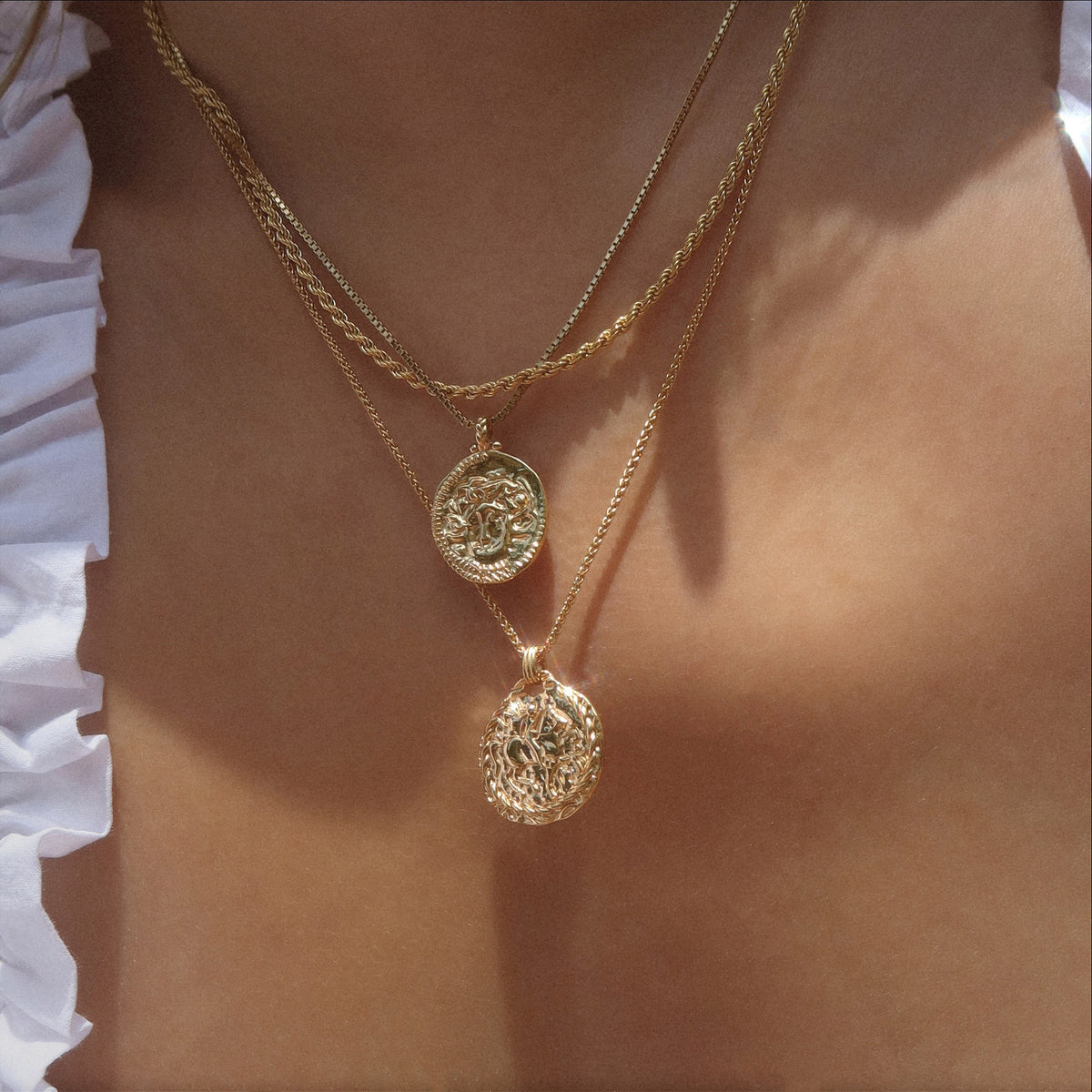 A close-up of a person wearing layered gold necklaces with ornate oval pendants, against sun-kissed skin and the edge of a white ruffled top visible on the side.