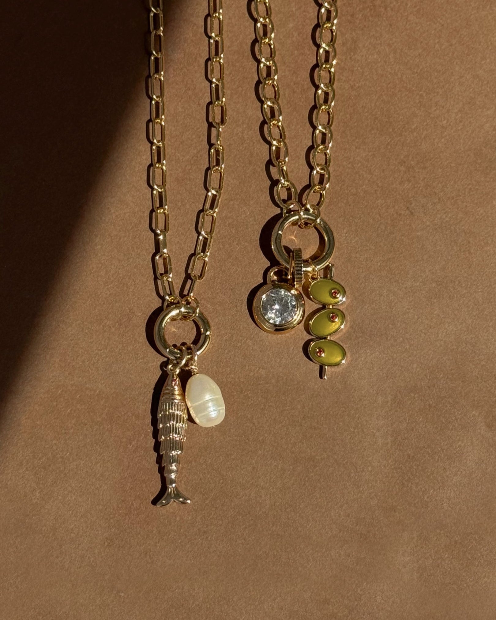 Two Darcy Charm Clip Necklaces by Mod + Jo on a neutral background: one with a paperclip chain, fish and pearl charms; the other with a clear gem and three small green bead charms.