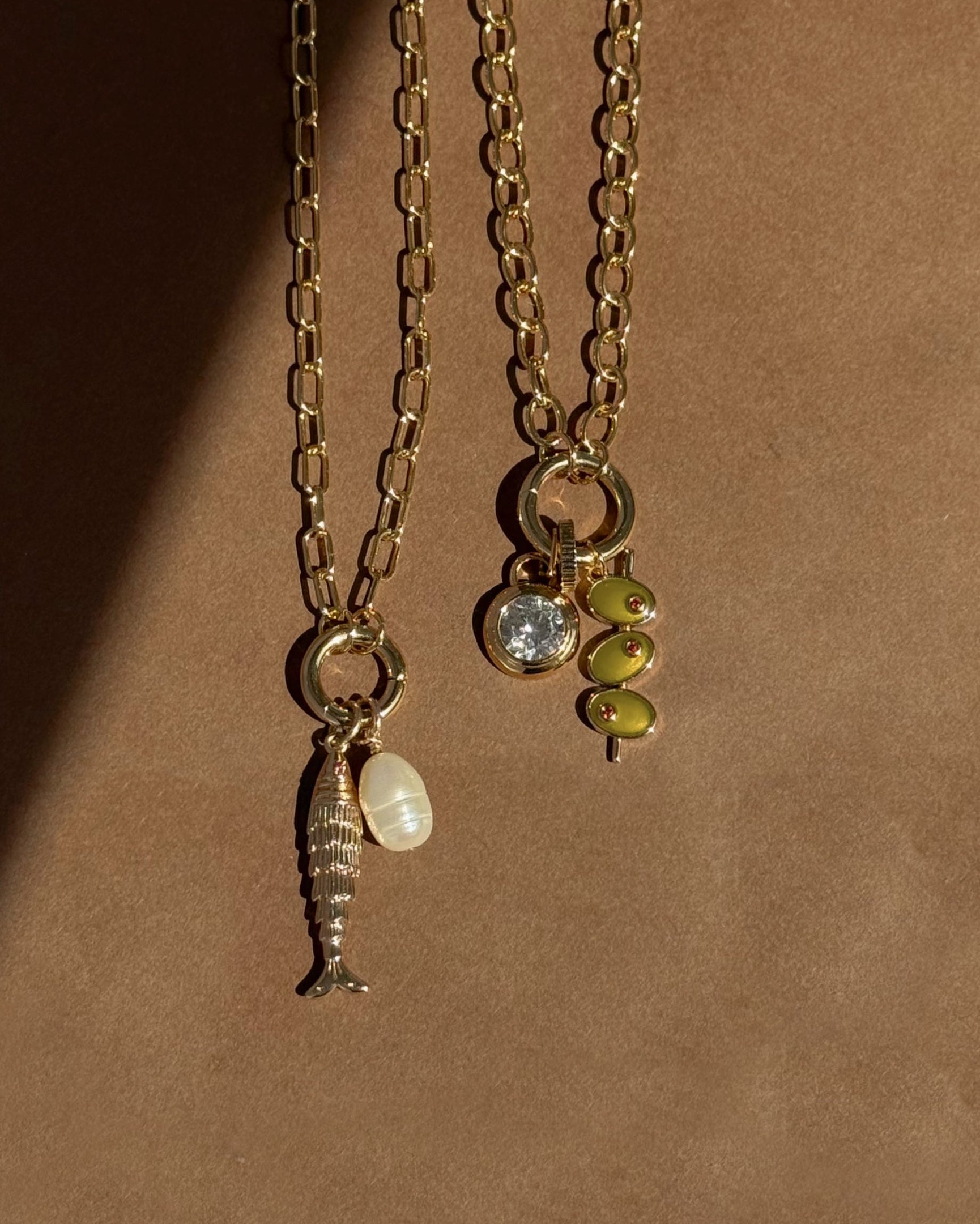 The Presley Charm Clip Necklace by Mod + Jo features two gold-plated chains: one with fish and pearl charms, the other with a round gem and green beads, both elegantly displayed on a brown surface.