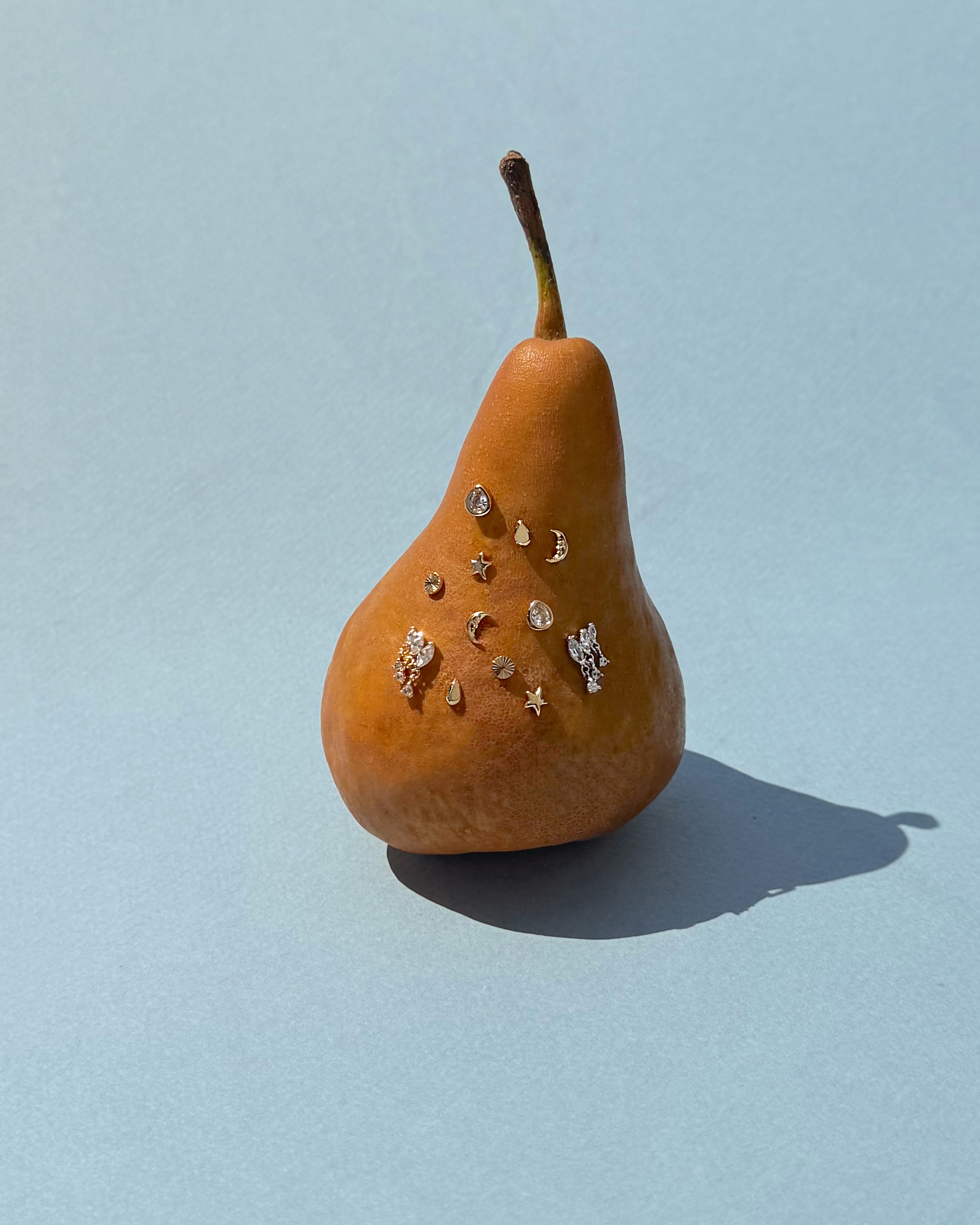 A brown pear stands upright on a light blue background, its front adorned with small, shiny charms shaped like stars, moons, and circles, reminiscent of the delicate gold plating of Mod + Jo’s Libbie Puffy Star Studs.