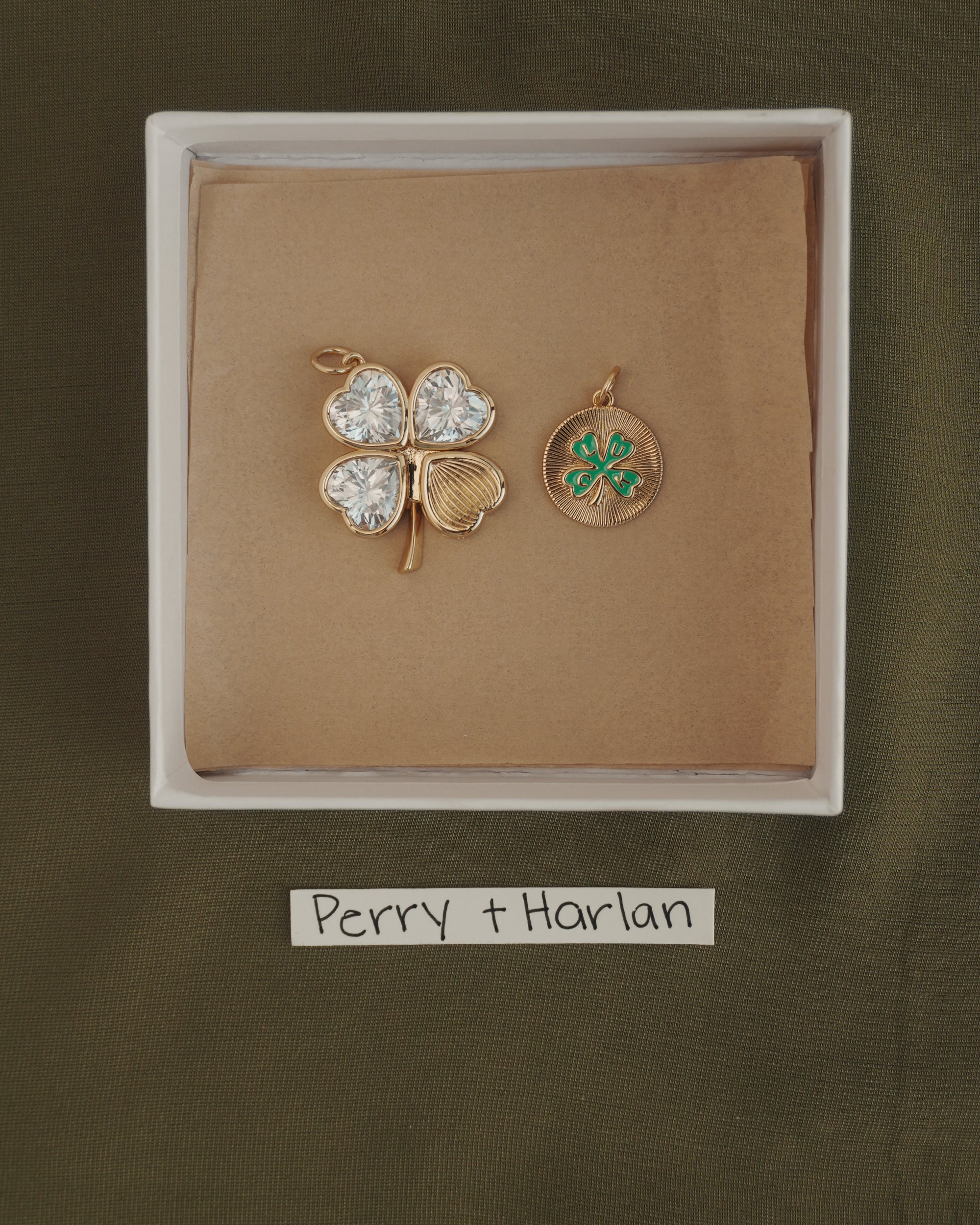 A white box displays the Perry Four-Leaf Clover Locket Charm by Mod + Jo—a gold locket in the shape of a four-leaf clover, its leaves shimmering with heart-cut CZ stones, paired with a small gold medallion adorned with a green clover. The strip below reads "Perry + Harlan.