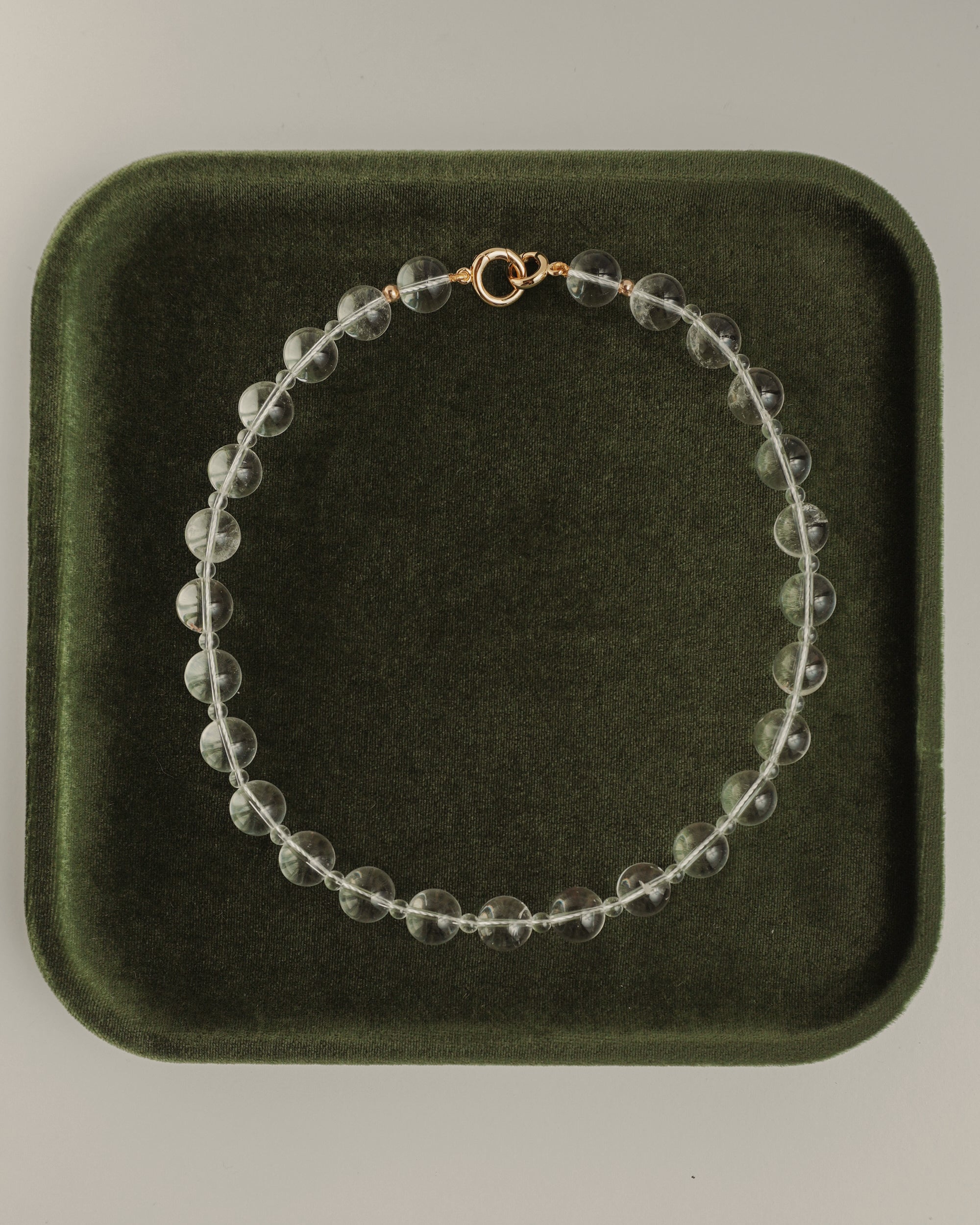 The Mod + Jo Portia Gemstone Necklace, featuring clear crystal quartz and a gold clasp, is displayed in a circle on a dark green velvet tray.