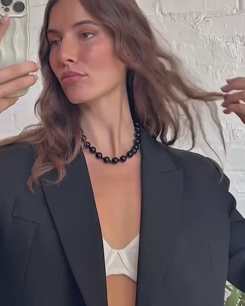 A woman taking a selfie video wearing a black blazer, a Mod + Jo Portia beaded necklace in black obsidian and zooming in on the necklace