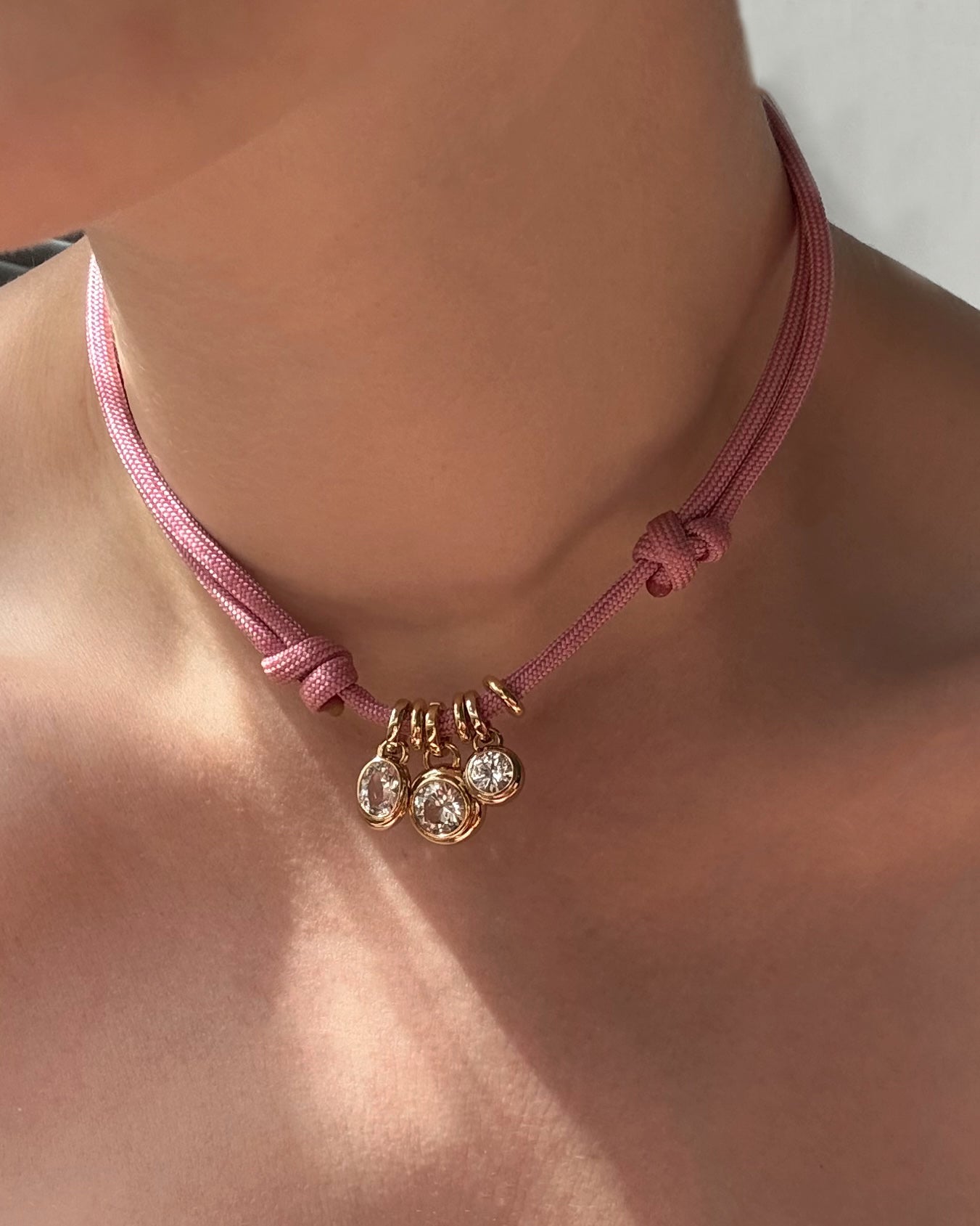 A close-up of a person's neck wearing Mod + Jo's *Preorder* Olly Sapphire Bezel Charm—a pink cord choker with side knots and three sparkling charms, including a lab-grown white sapphire set in 14k gold at the center.