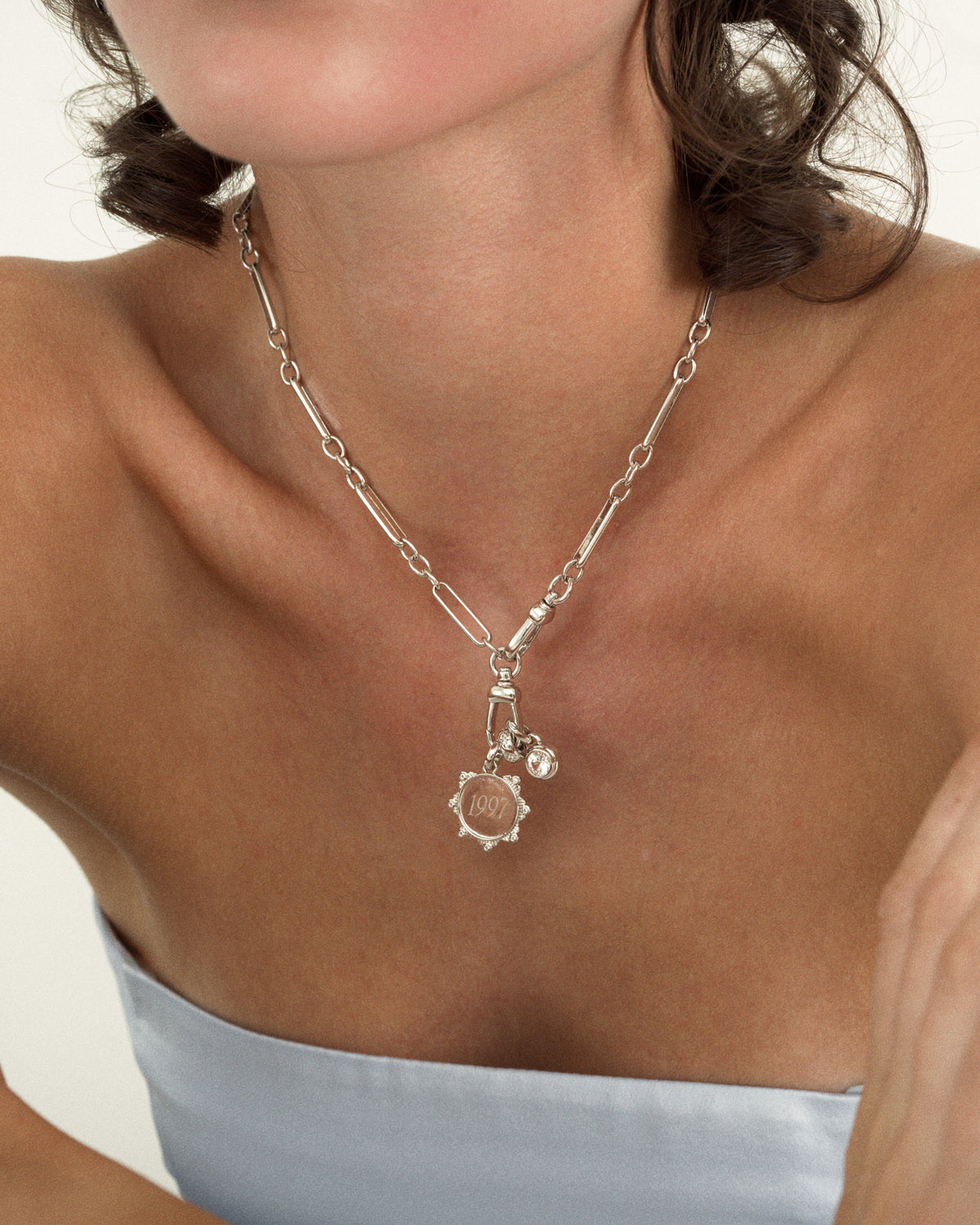 The Mod + Jo Remi Engravable Charm is a silver-plated chain necklace featuring a circular charm engraved with the number 1997 and a small round cubic zirconia crystal charm.