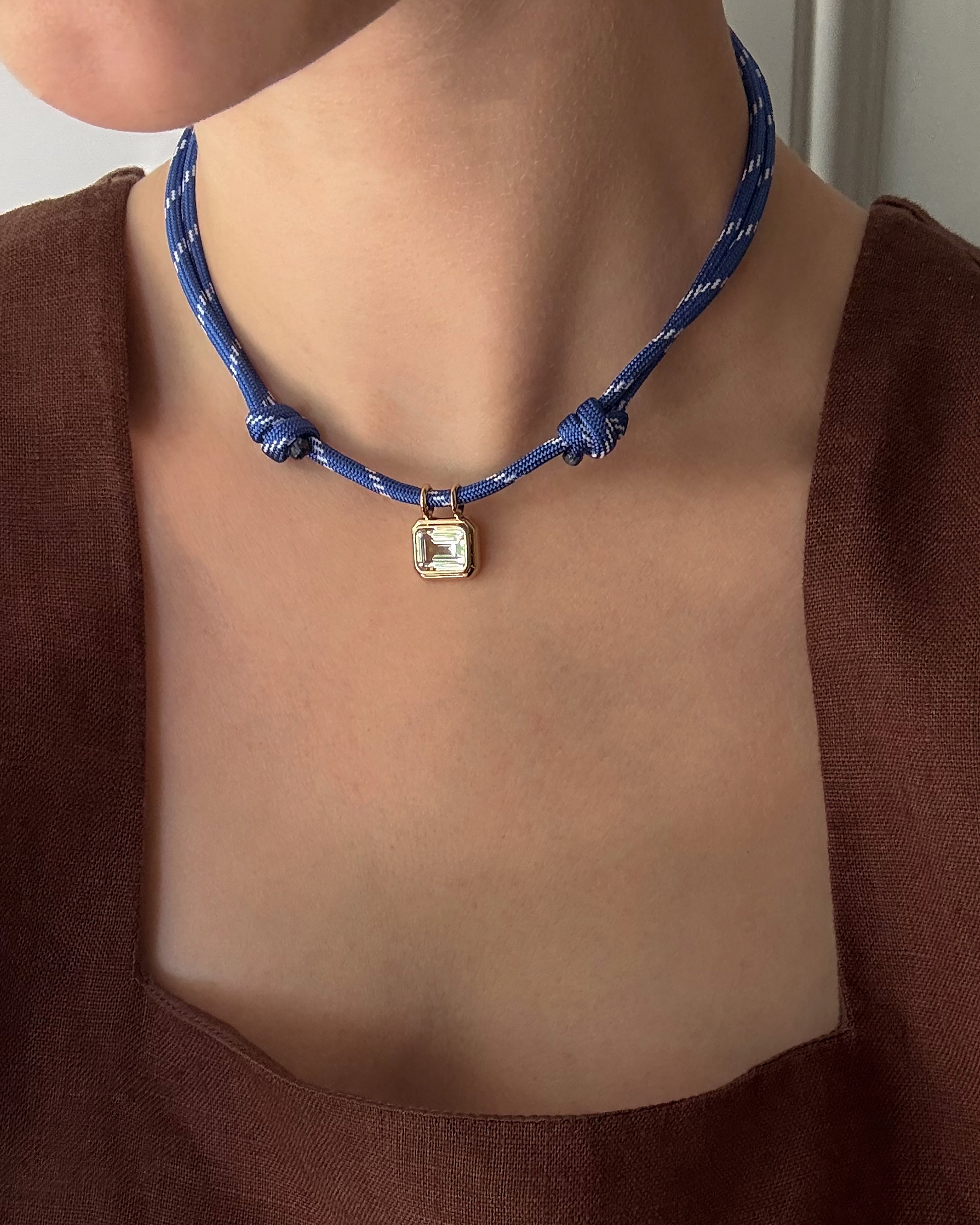 A person wearing the Mod + Jo Elsie Sapphire Bezel Charm—a gold-framed, square, lab-grown white sapphire pendant—on a blue cord choker with two knots. They are dressed in a brown, square-neck top.