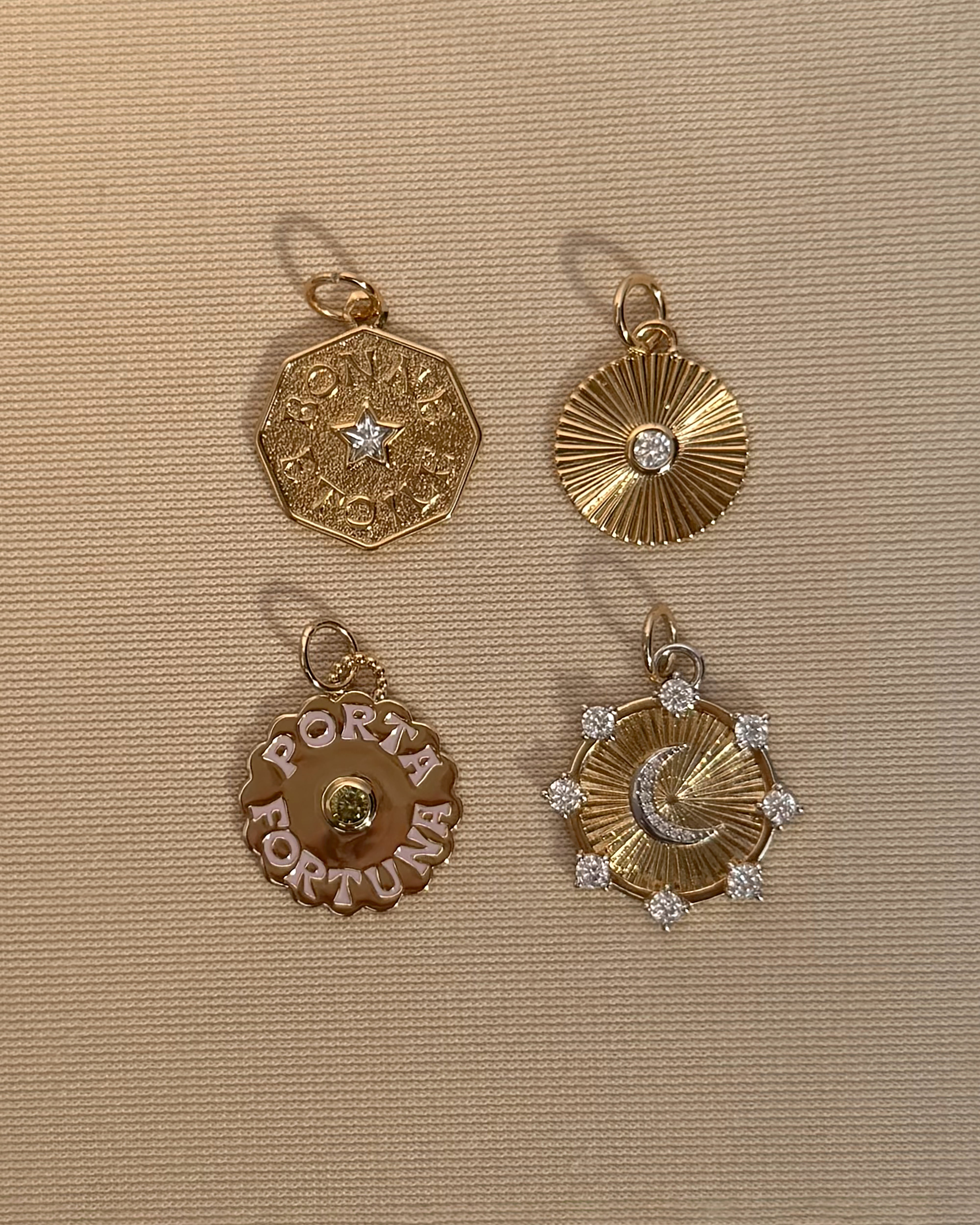 Four gold pendants from Mod + Jo are displayed on a beige textured background: an octagon with a star, a sunburst pendant with a gemstone reminiscent of a lucky charm, the "Porta Fortuna Charm" in 14k gold, and a sun-and-moon design adorned with small gemstones.