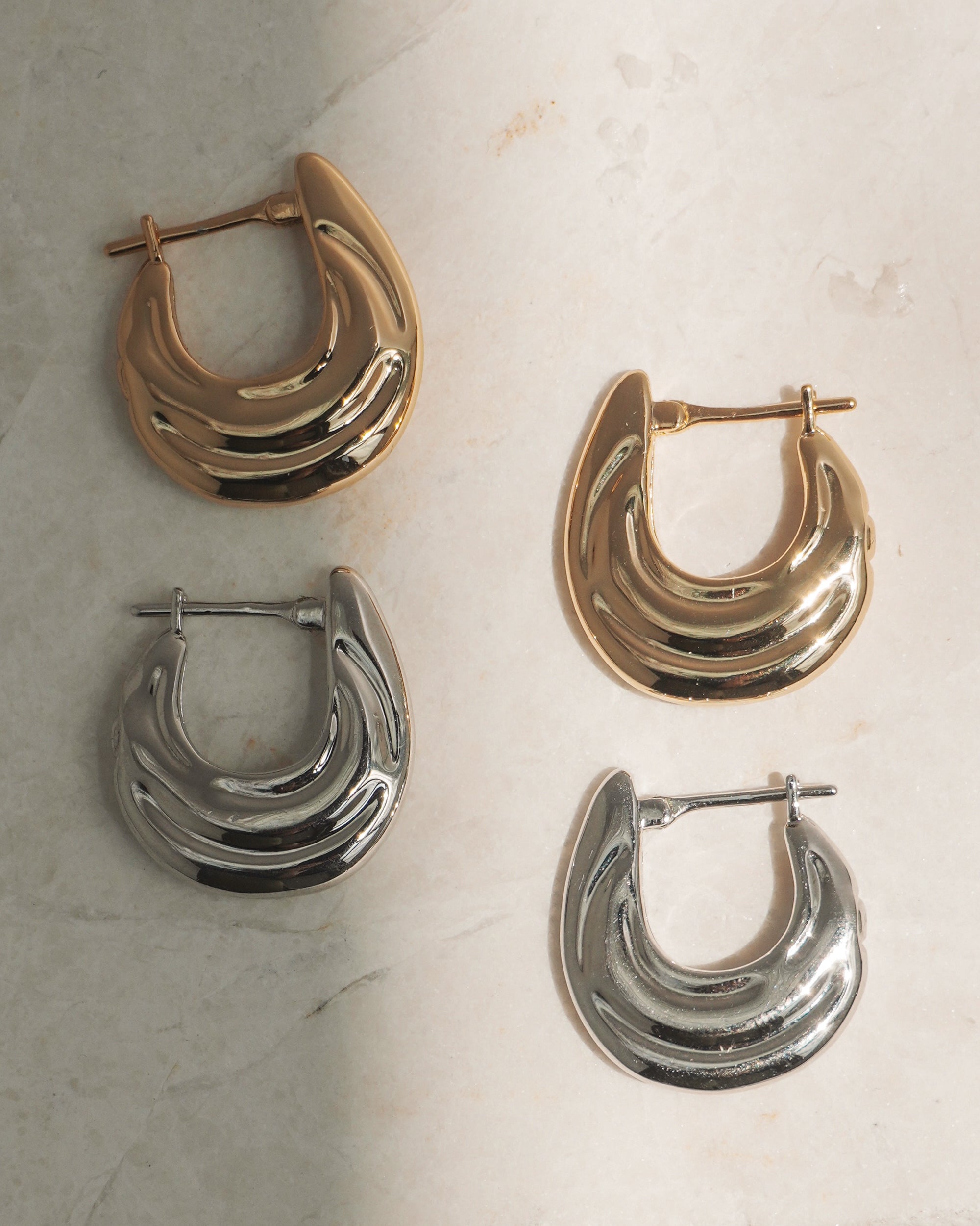 Two pairs of Samara Hoops by Mod + Jo, one gold and one silver, rest on a marble surface. Each features a crescent-shaped, layered design with advanced plating and secure latch back closures.