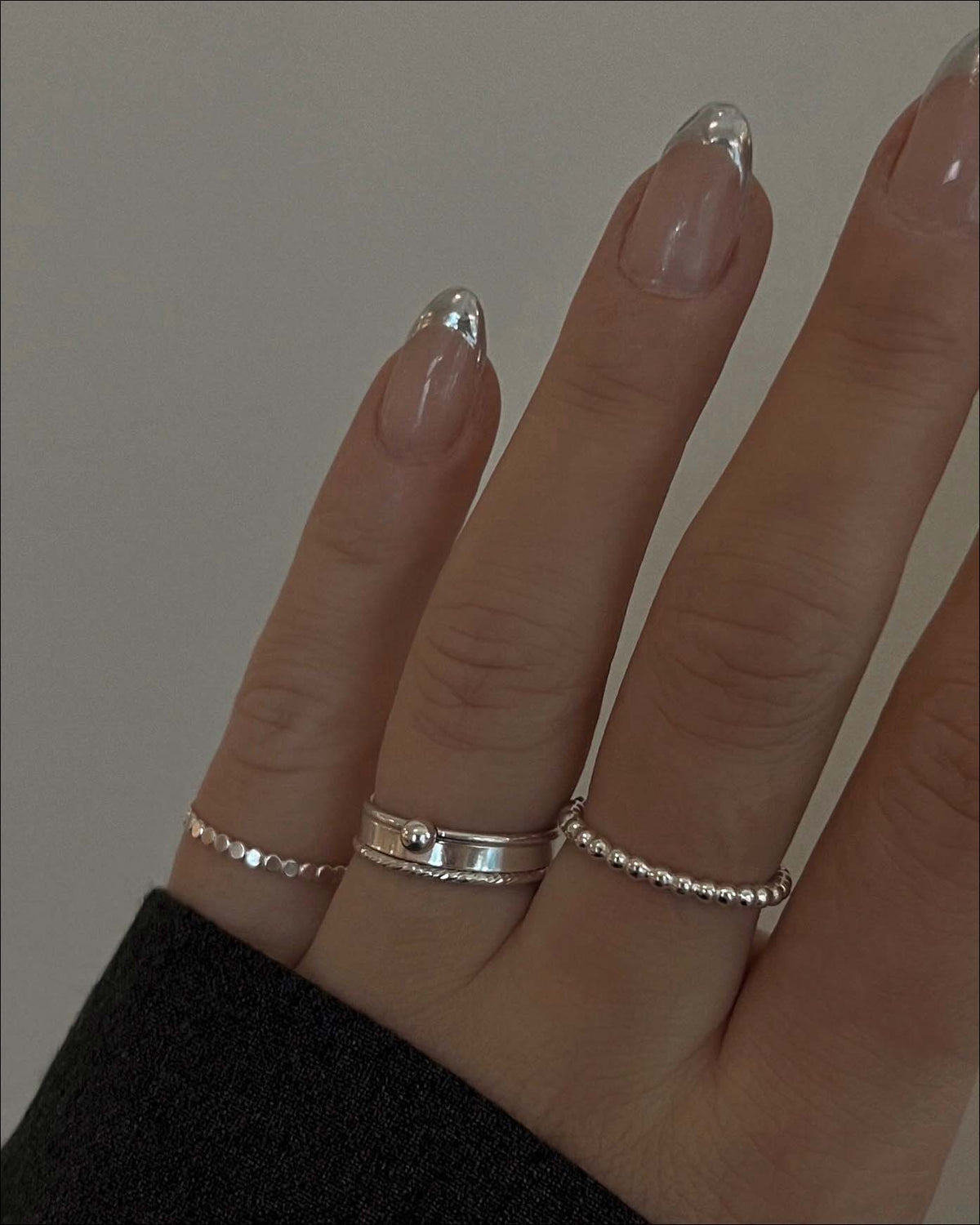 A hand with almond-shaped, clear-tipped nails wears three silver rings: a thin beaded band, a double band with a small sphere accent, and another thin beaded band on different fingers.