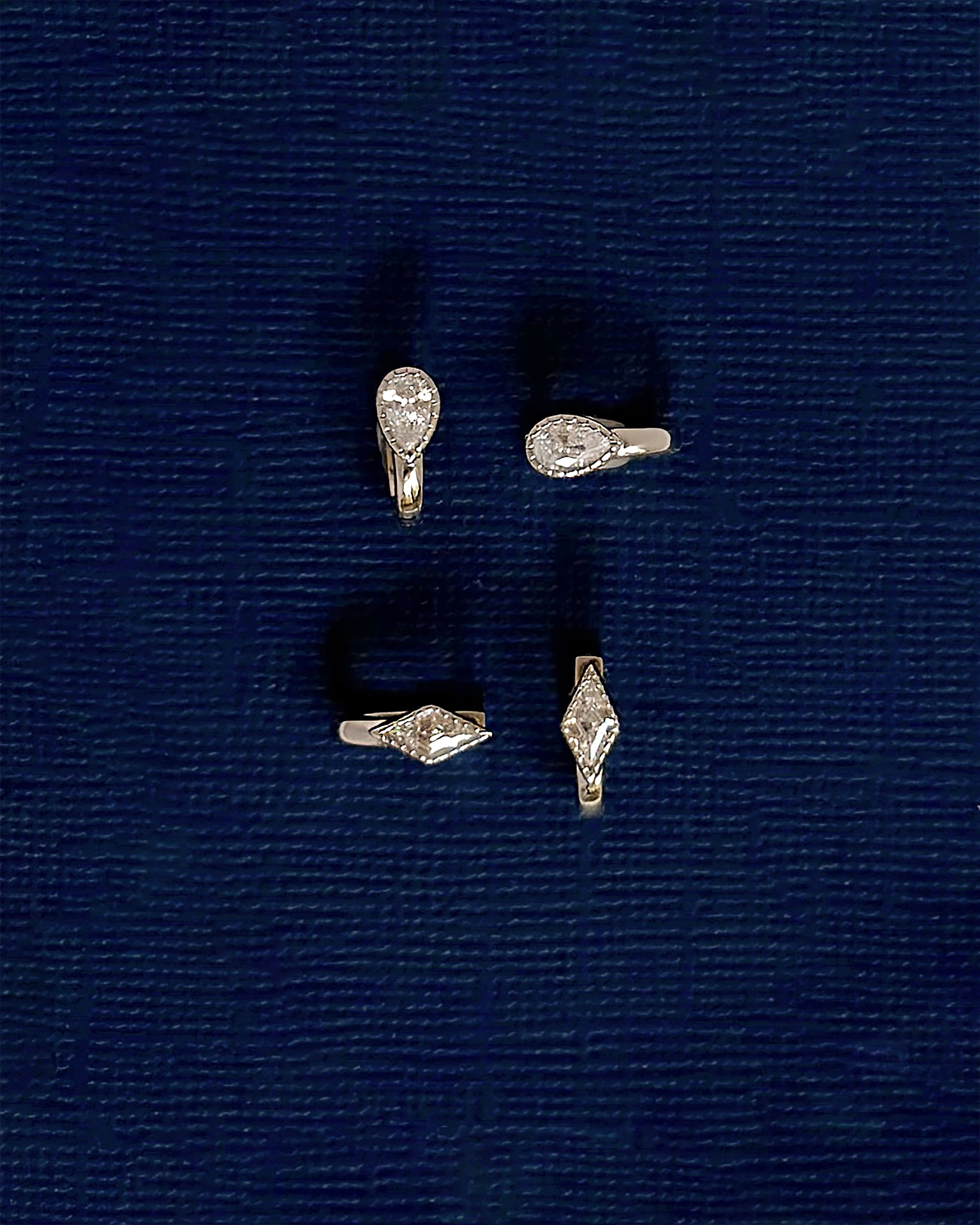 Four Phoebe Kite Huggies by Mod + Jo, featuring diamond-shaped and teardrop-shaped clear stones accented with dazzling kite-cut cubic zirconia, are arranged in a square on a textured dark blue surface.