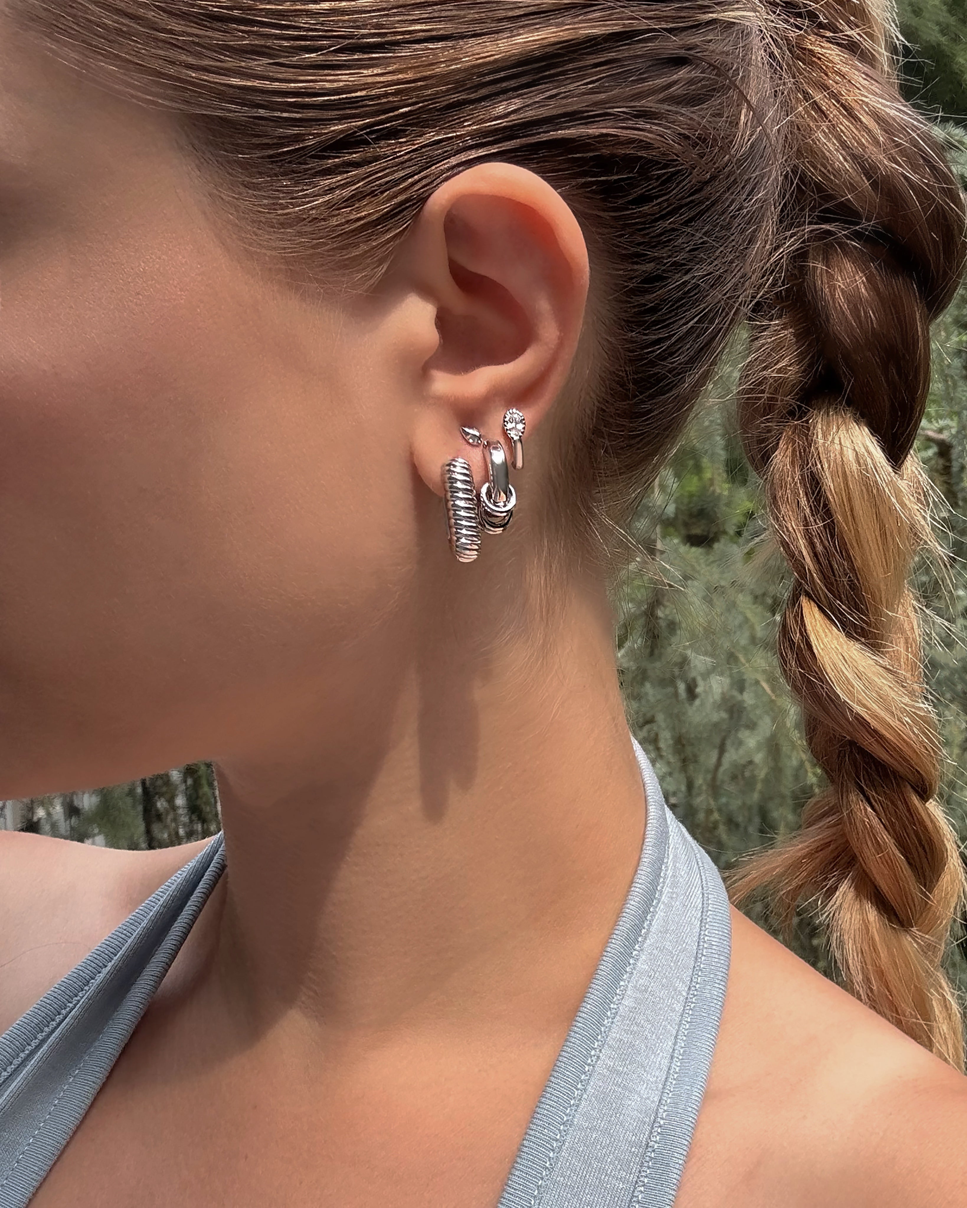 A woman with a braided ponytail wears the Mod + Jo Elly French Rope Hoops. She is dressed in a light gray halter top and stands outdoors with greenery softly blurred in the background.