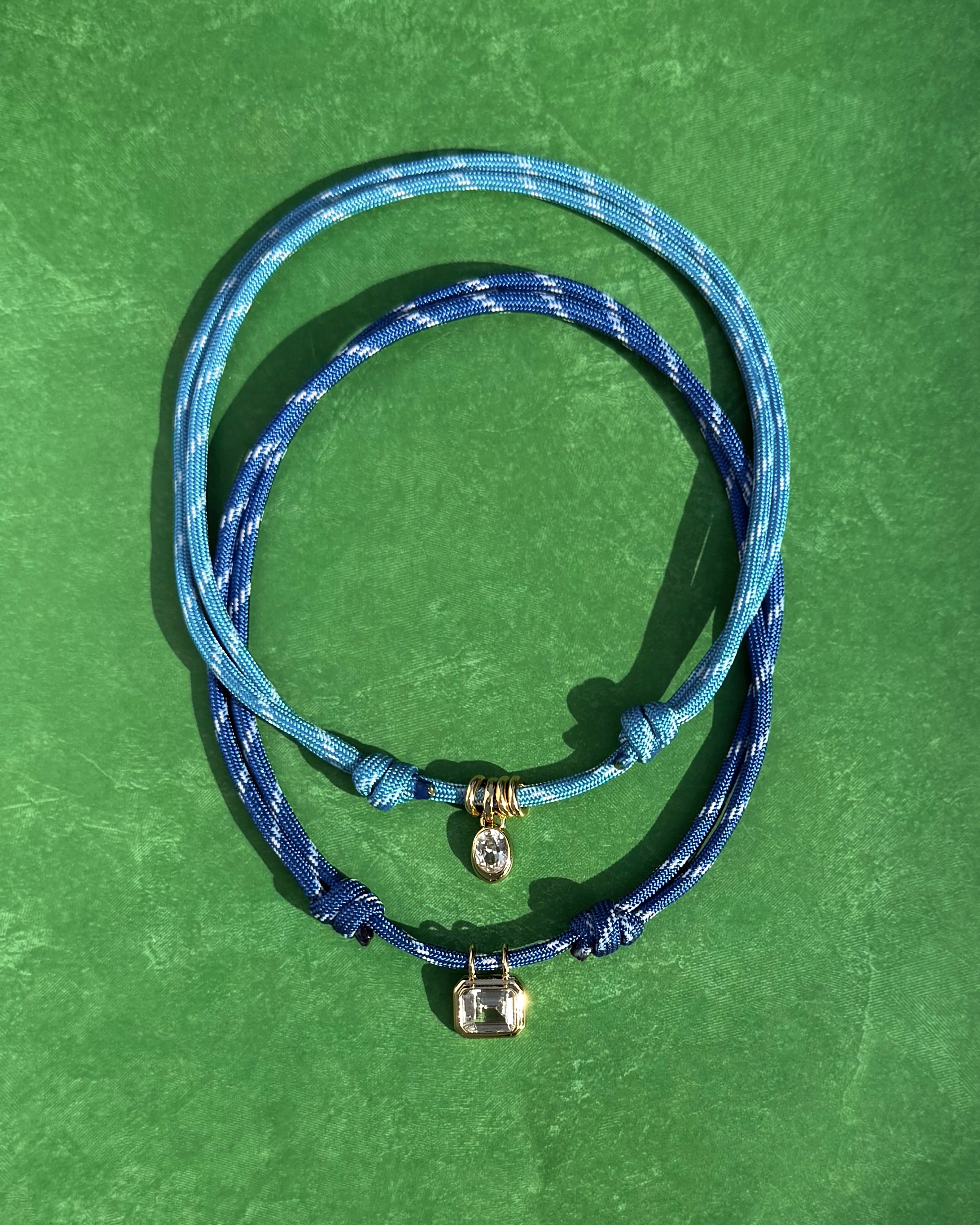 Two adjustable blue cord bracelets, one with a silver disc charm and gem, the other with a silver padlock, rest on green. Pair them with the *Preorder* Tommy Sapphire Pendant Necklace by Mod + Jo for a chic layered look.