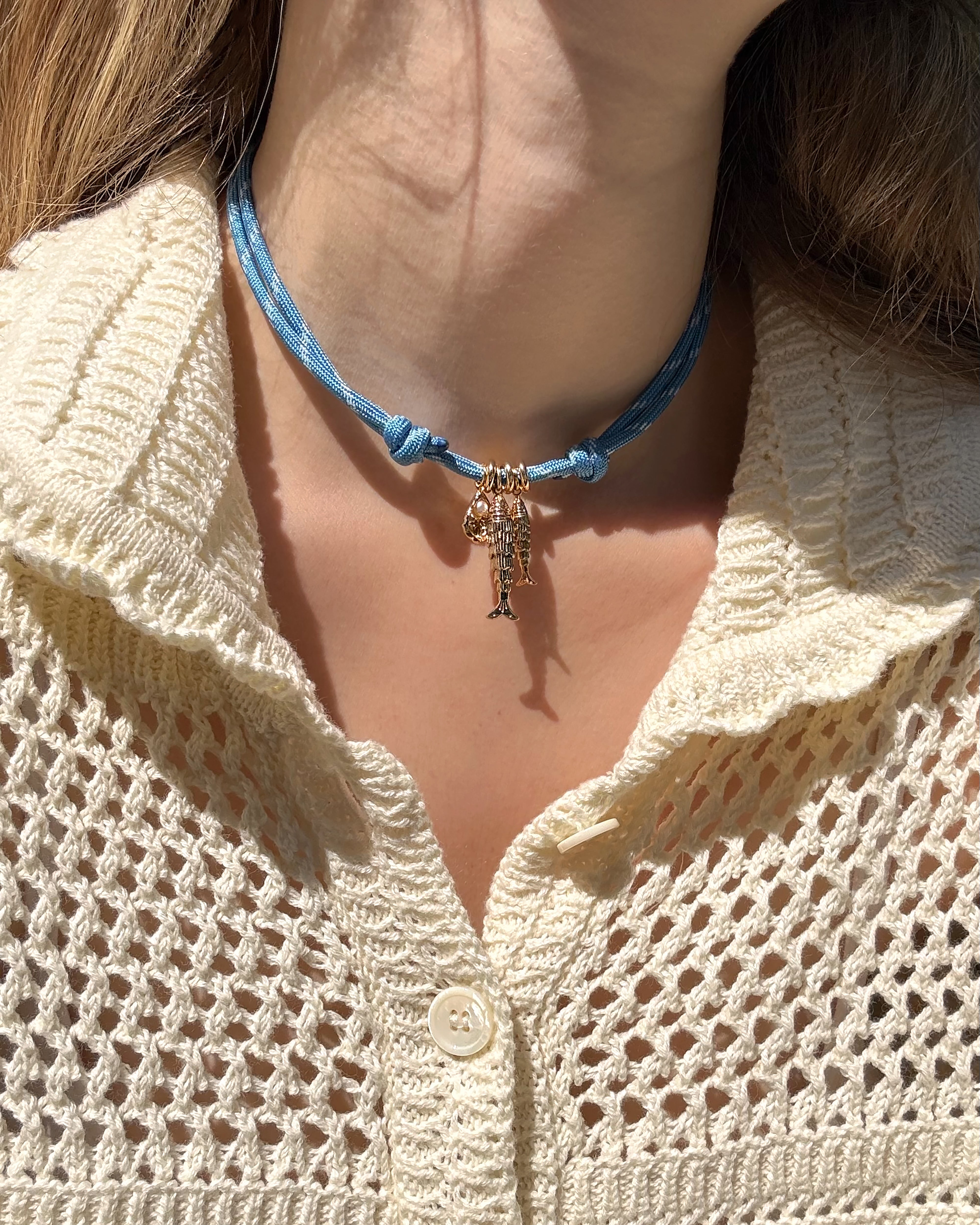 A person wearing a cream knit sweater with the *Preorder* Tommy Cord Necklace by Mod + Jo, featuring gold-plated spacers and gold-toned saxophone and musical note charms. Sunlight casts shadows across their neck and collar.
