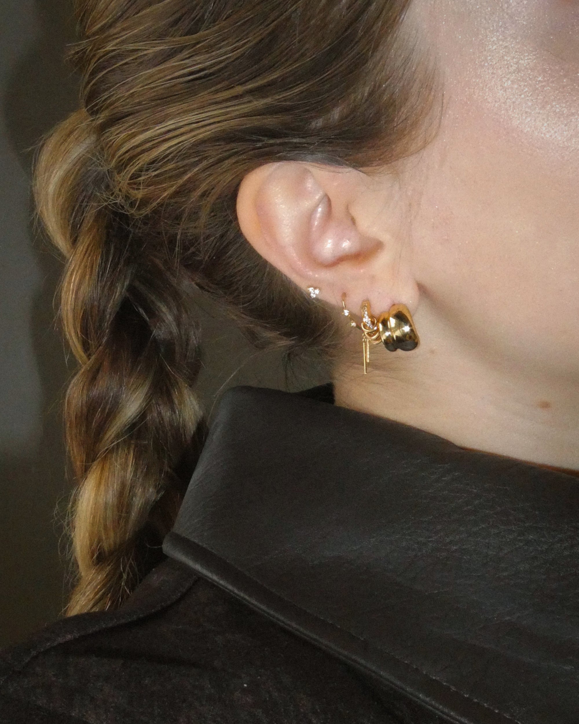 Close-up of a person’s ear adorned with Mod + Jo’s Petite Sofie Volume Hoops, featuring multiple gold hoops and a small stud. Brown hair is styled in a braid and the person wears a dark, textured jacket with a wide collar.