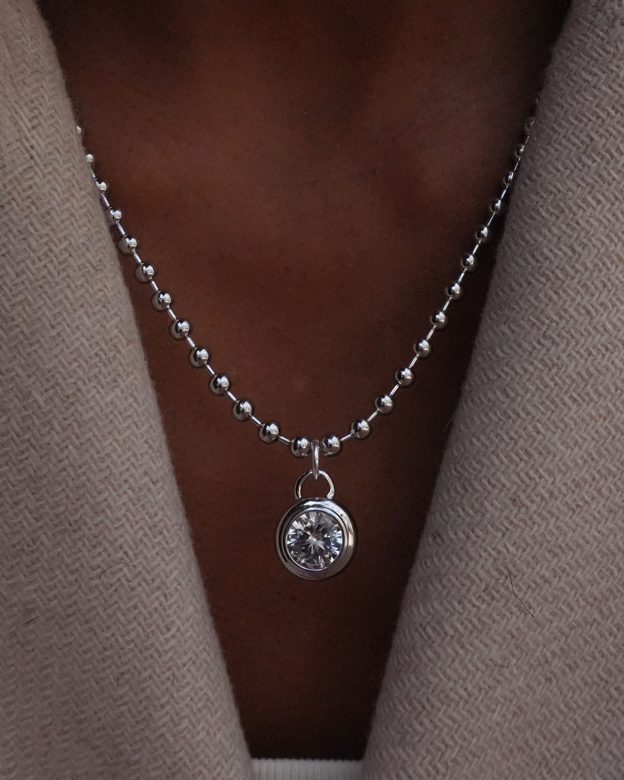 Silver ball chain necklace with a round sapphire bezel pendant on a person wearing a beige cardigan.