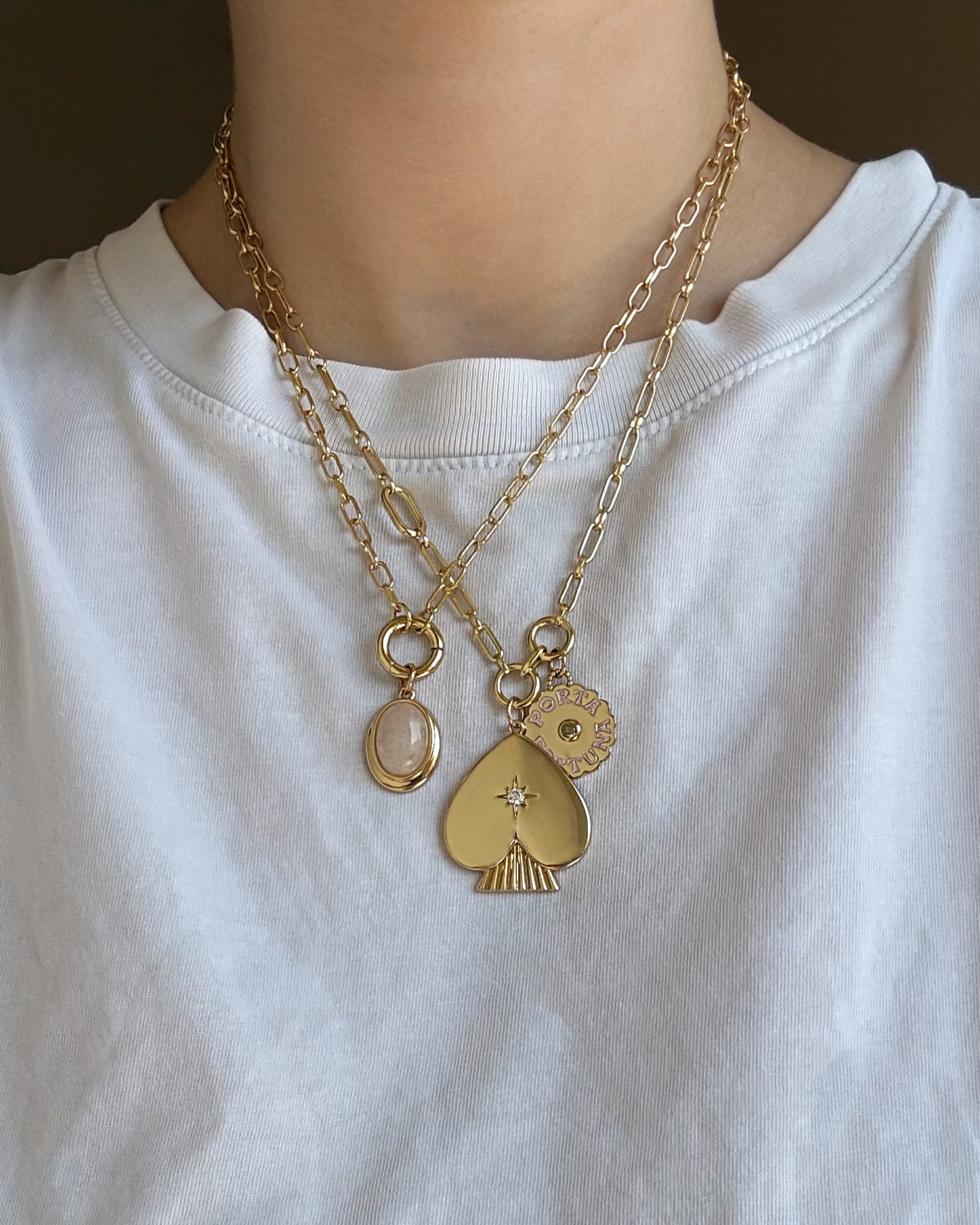 A person wearing a white shirt with two layered 14k gold chain necklaces from Mod + Jo, each adorned with distinctive pendants: a small oval, a spade with a star, and the Rory Gemstone Charm—a round sun-shaped piece that embodies self-love.