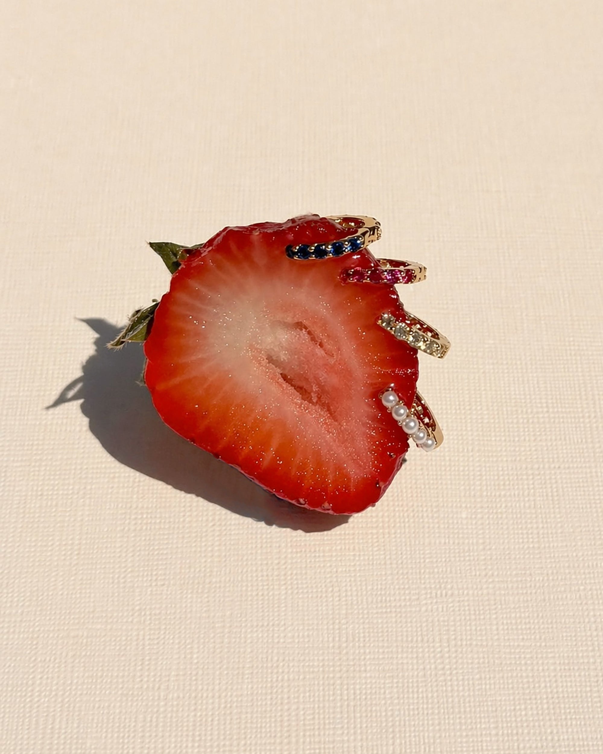 A sliced strawberry is pinned with several Mod + Jo Harlow Reversible Birthstone Huggies, gold rings adorned with colorful gemstones and pearls, all arranged on a light textured background.