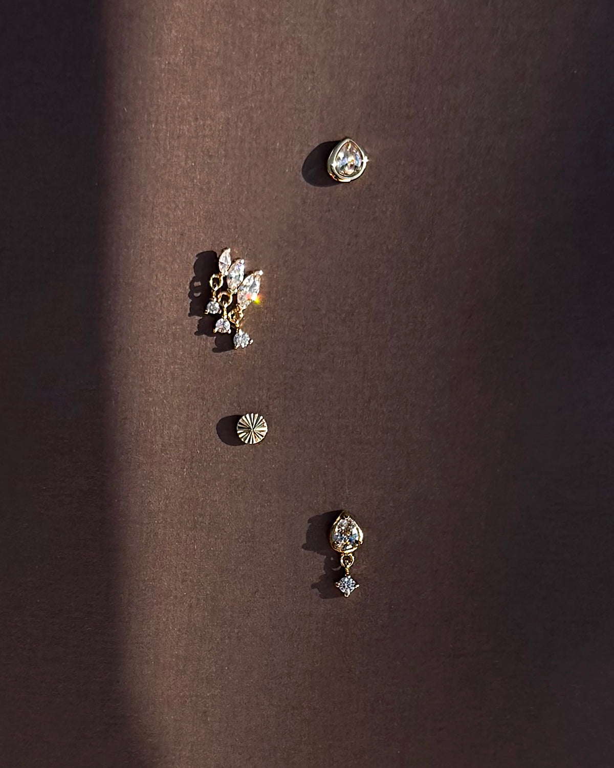 Four unique earrings from Mod + Jo, including the Joni Bezel Studs, are arranged vertically on a dark brown background. They showcase gold- and silver-toned designs in various shapes with sparkling stones like cubic zirconia, all casting soft shadows.