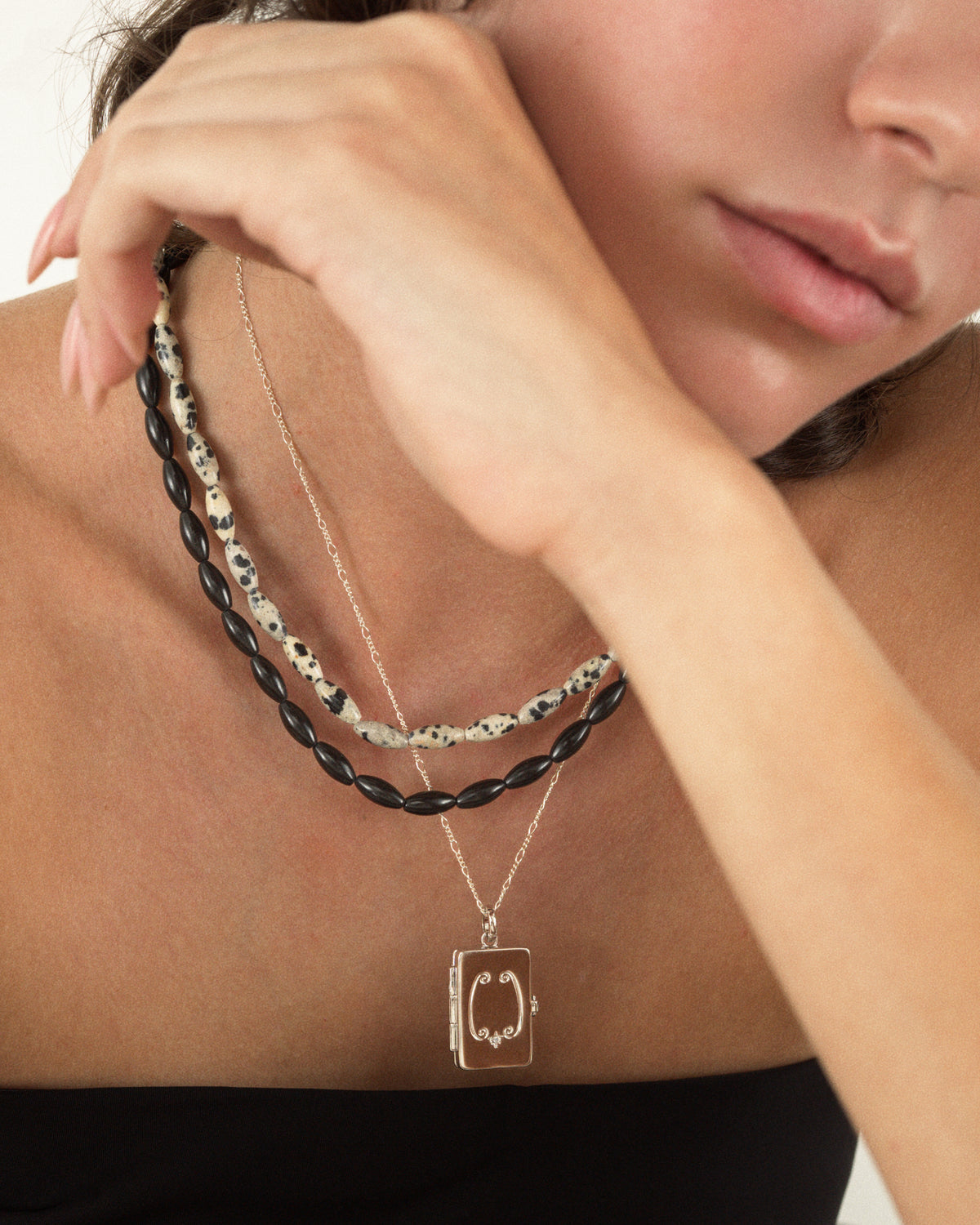 A person wearing a Sutton Gemstone Necklace layered with two other necklaces: one with black beads and a delicate silver chain with a rectangular locket pendant.