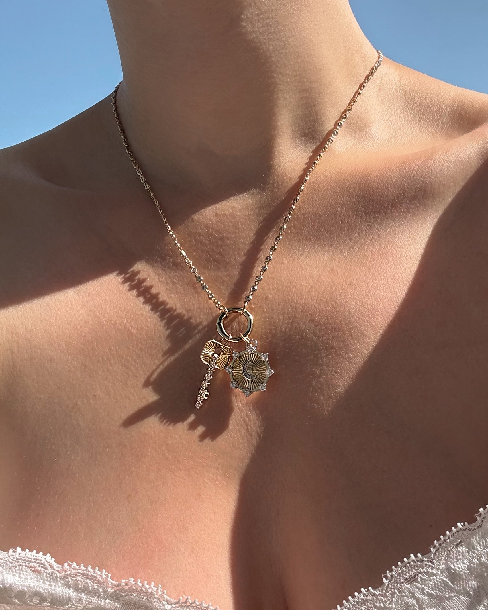 A close-up of a person’s neck wearing Mod + Jo’s Noah Key Charm—a gold plated necklace adorned with key and flower-shaped charms—against a clear blue sky and dressed in white lace, showcasing an elegant piece of symbolic jewelry that beautifully blends style and meaning.