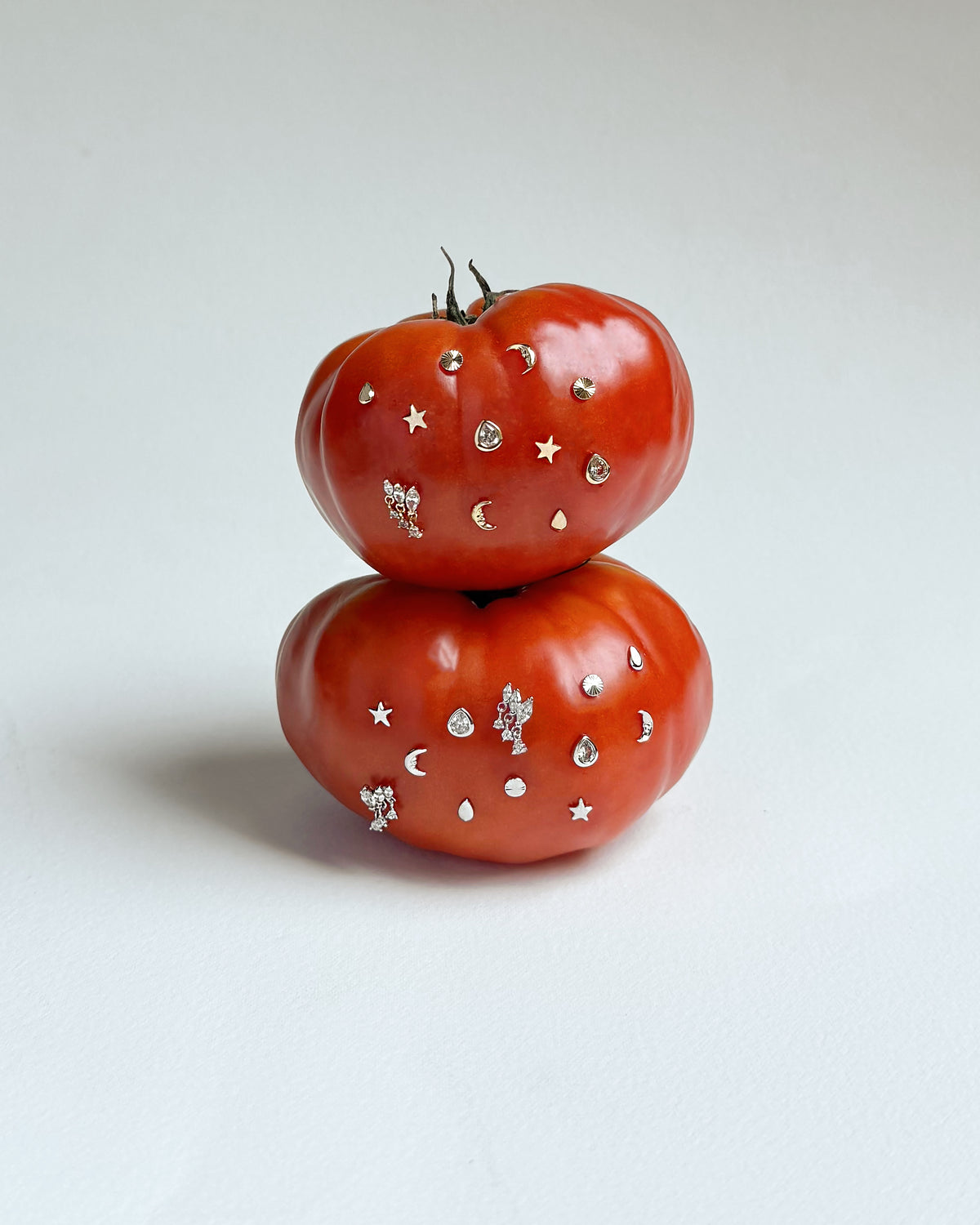 Two red tomatoes stacked vertically, each decorated with an assortment of Mod + Jo Celestial Studs Set earrings, including moon and star designs.