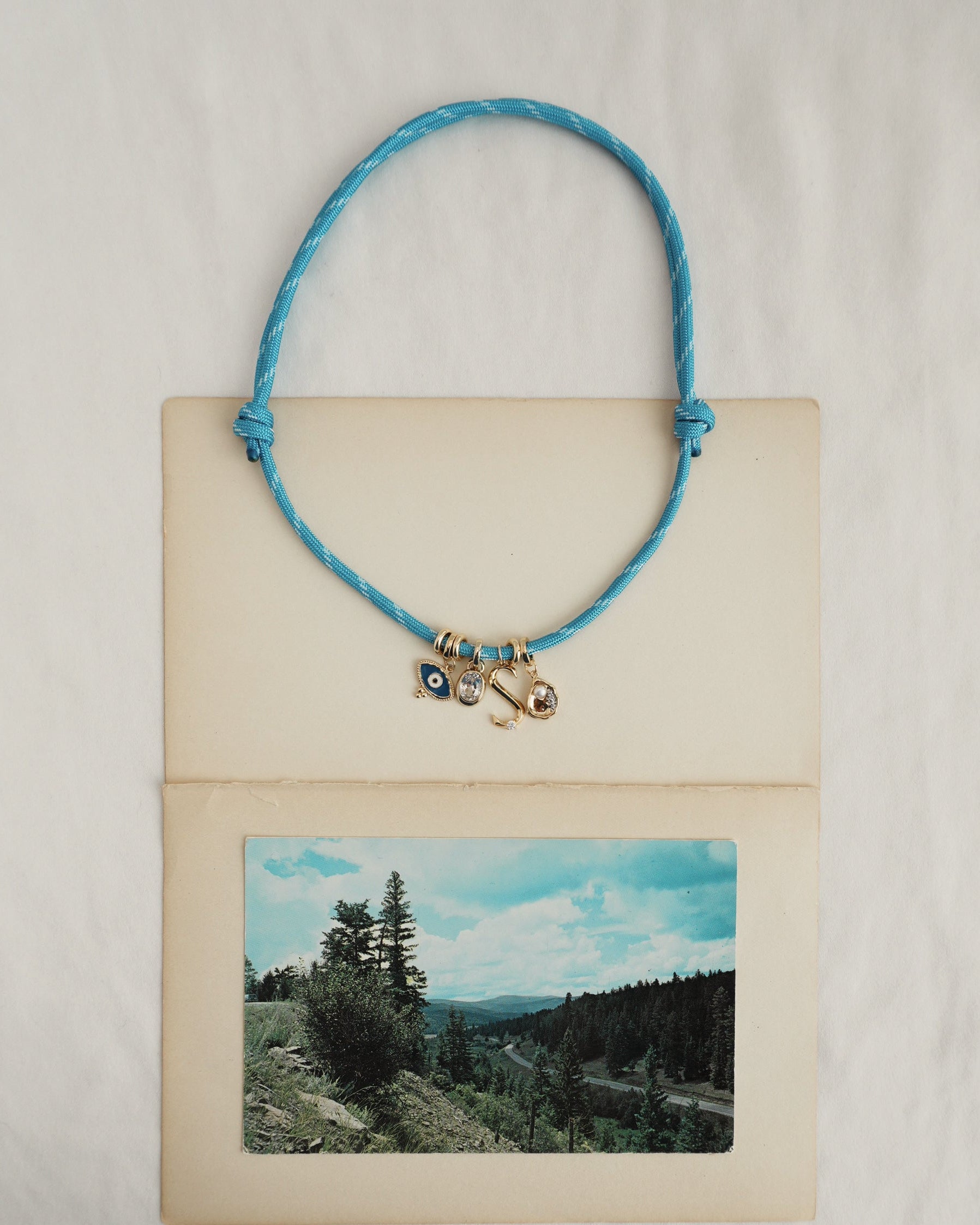 The *Preorder* Tommy Cord Necklace by Mod + Jo, featuring gold plated spacers and small charms, rests on an open vintage photo album atop white fabric. A landscape photograph with trees and hills is visible beneath the necklace.